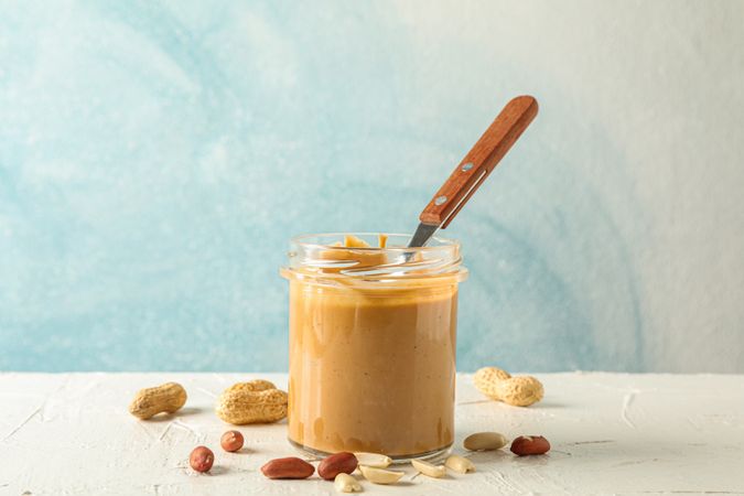 Glass jar with creamy peanut butter and spoon, and peanut on table against light background, space for text