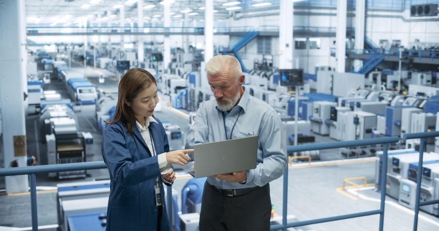 Factory Managers Working on Laptop at a Modern Manufacture with Autonomous Robot Hands. Female and Male Specialists Monitoring Technical Data on a Computer, Analyzing Reports and Planning Updates