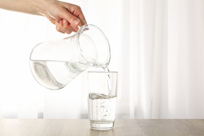 Hand pouring water in glass from pitcher in bright room with curtain