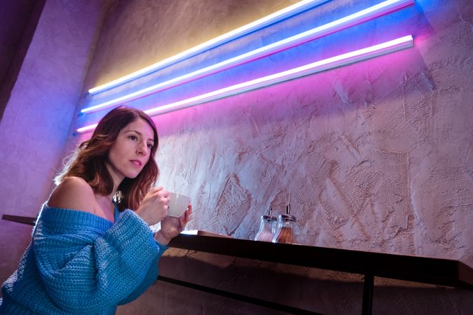 Positive woman with cup at table near wall with neon lamps
