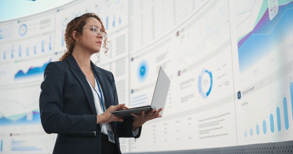 Successful Hispanic Businesswoman Holding Laptop Computer And Reviewing Financial Reports On Digital Screen In Monitoring Office.