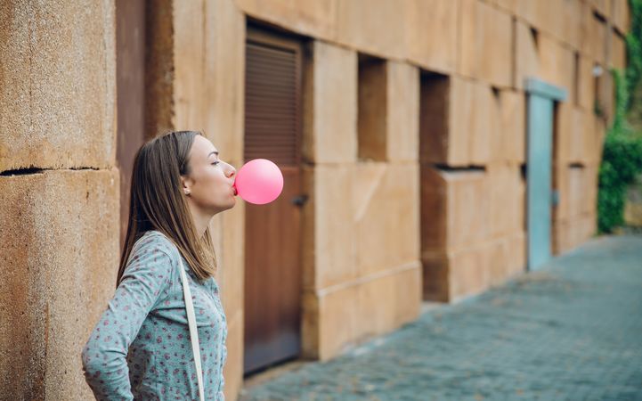 Side view of woman standing outside in front of stone wall blowing bubble gum with space for text