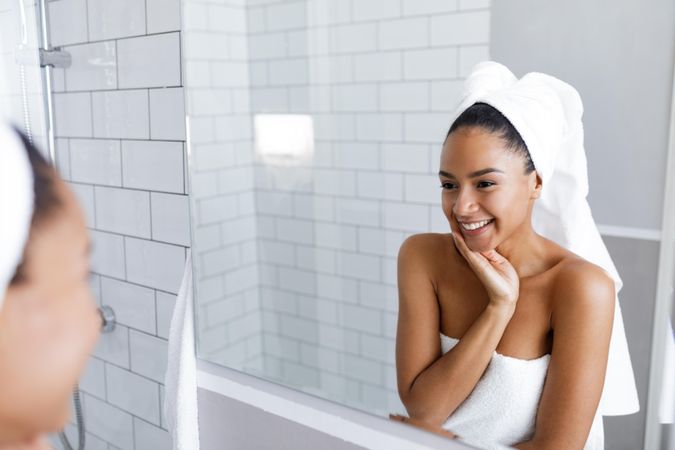 Smiling woman in towel touching her jaw while looking in the mirror