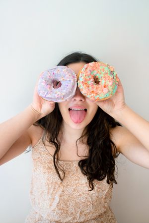 Playful woman making glasses of donuts while standing by white wall
