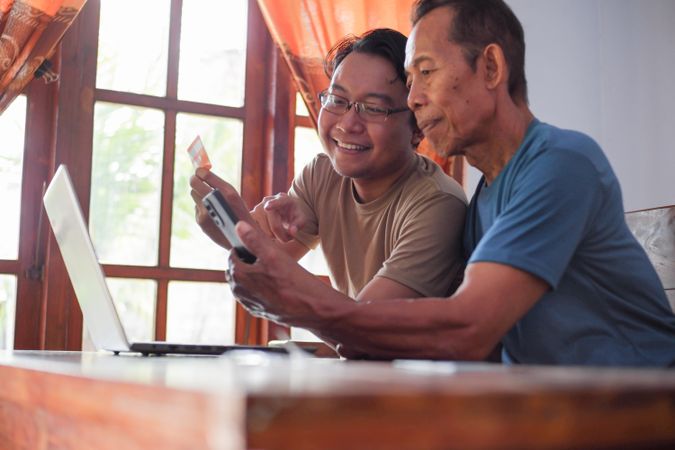 Happy adult son and mature father pay online using laptop and credit card