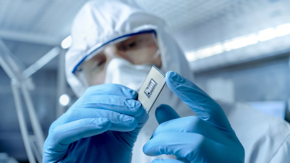 In Ultra Modern Electronic Manufacturing Factory Design Engineer in Sterile Coverall Holds Microchip with Gloves and Examines it.