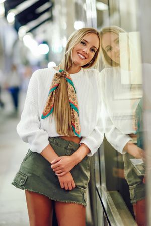 Chic happy woman with scarf in hair leaning on glass of shop outside
