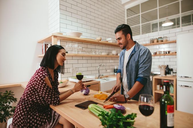 Young woman drinking wine while husband chops vegetables on kitchen counter