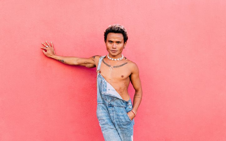 Male in open overalls looking at camera leaning against a pink wall