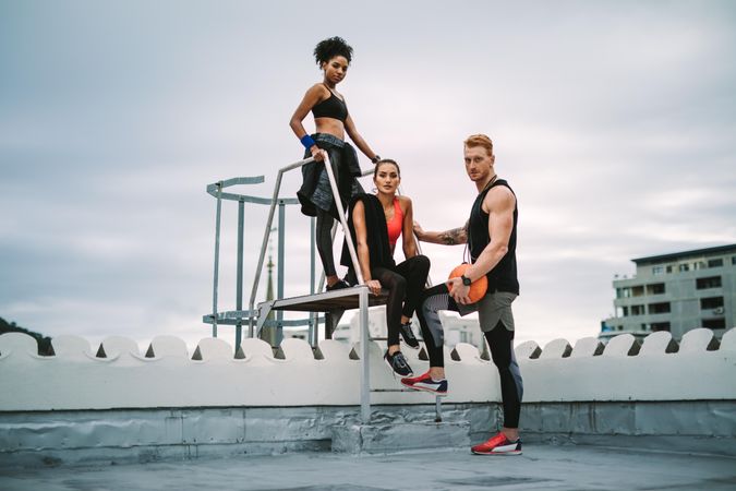Two fitness women with a man relaxing after workout on rooftop standing near the stairs