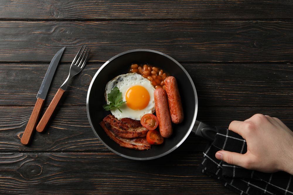 Top view of hand reaching for pan with fried breakfast of eggs, bacon and sausage