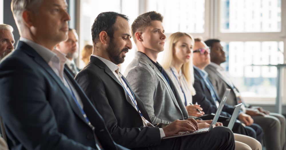 Multiethnic Man Sitting in Crowded Auditorium at Conference. Middle Eastern Businessman Listening To Keynote, Typing On Laptop. Specialist Watching Technology Presentation About Software Solutions.