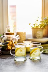 Healthy beverage with lemon and ginger in glass tea cups at kitchen table with mugs, candle and herbs at window background with natural light. Warming hot drinks with citrus fruit. Front view.