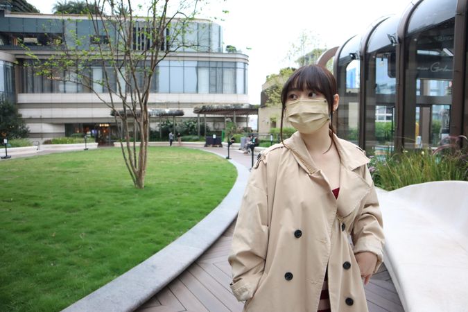 East Asian woman in beige coat with facemask standing near building