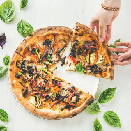 Quartered wood fired vegetable pizza with hand taking slice, square crop