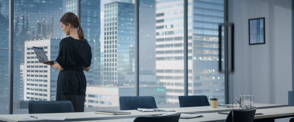 Businesswoman Sitting Down At Her Desk And Starts Typing On Laptop Computer In Corner Office With Megapolis Cityview.