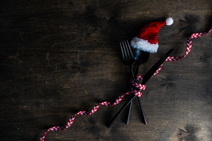 Cutlery setting with red Santa hat and ribbon on silverware on rustic background