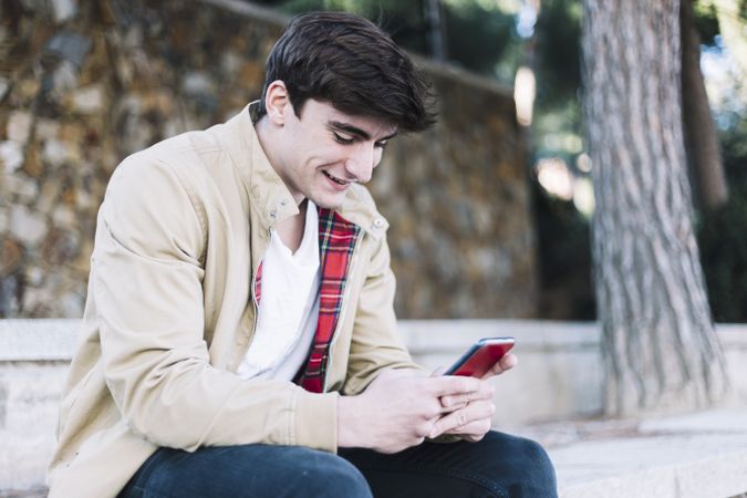 Young man using mobile phone and smiling while sitting outdoors