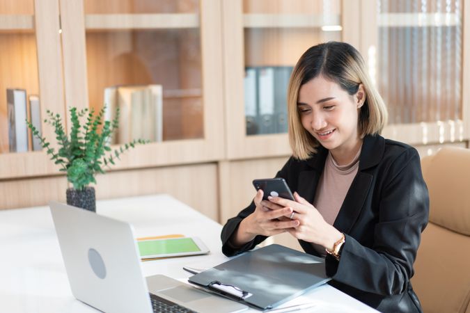 Woman smiling at phone while sitting at her desk with laptop