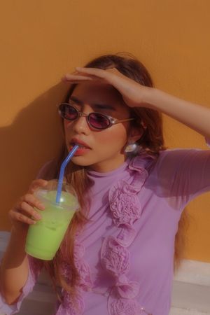 Young woman in purple top with sunglasses drinking green juice blocking sun with hand