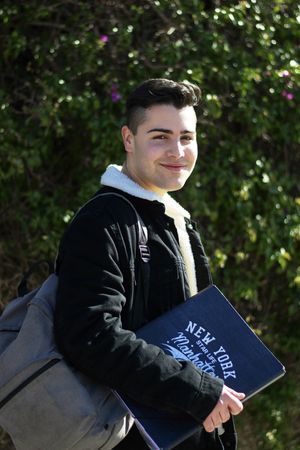 Young student holding notebook and backpack while smiling on camera