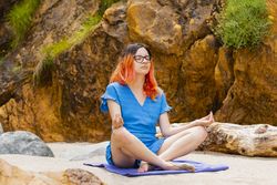 A young, orange-haired woman practices yoga and meditates on the beach in summer
