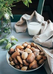 Glass of almond milk next to plate of almonds in shells on kitchen table