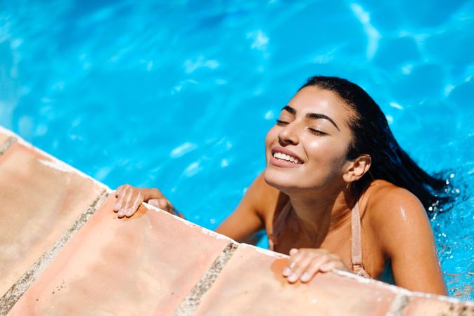 Beautiful Arab woman grasping side of swimming pool with eyes closed and head up