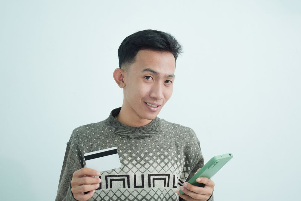 Curious Asian businessman in suit holding blank credit card and smartphone
