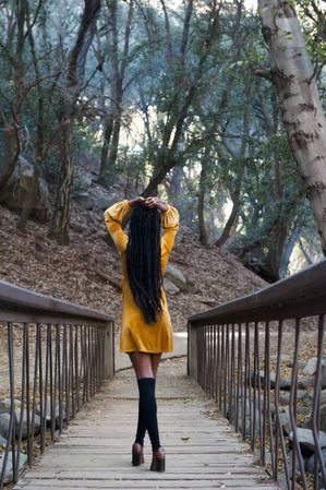Woman in yellow dress walking on a bridge facing away from the camera with hands above her head