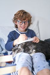 Cheerful boy doing homework on bed with dog