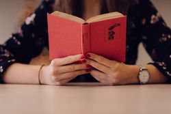 Cropped image of woman sitting at a table reading a book with red cover 0vKOR5