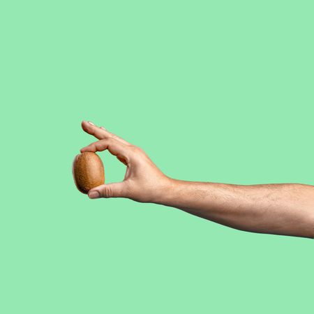 Hand holding a kiwi fruit on a mint green background.