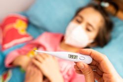 Girl sick in bed wearing face mask with adult taking her temperature