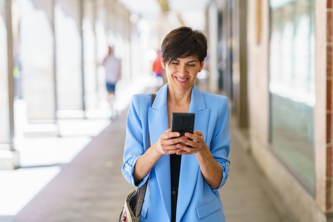 Smiling woman in blue blazer checking phone while walking down street