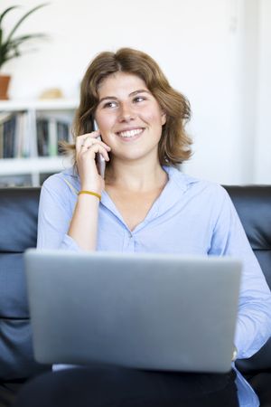 Businesswoman sitting on sofa at home using a laptop and chatting on phone