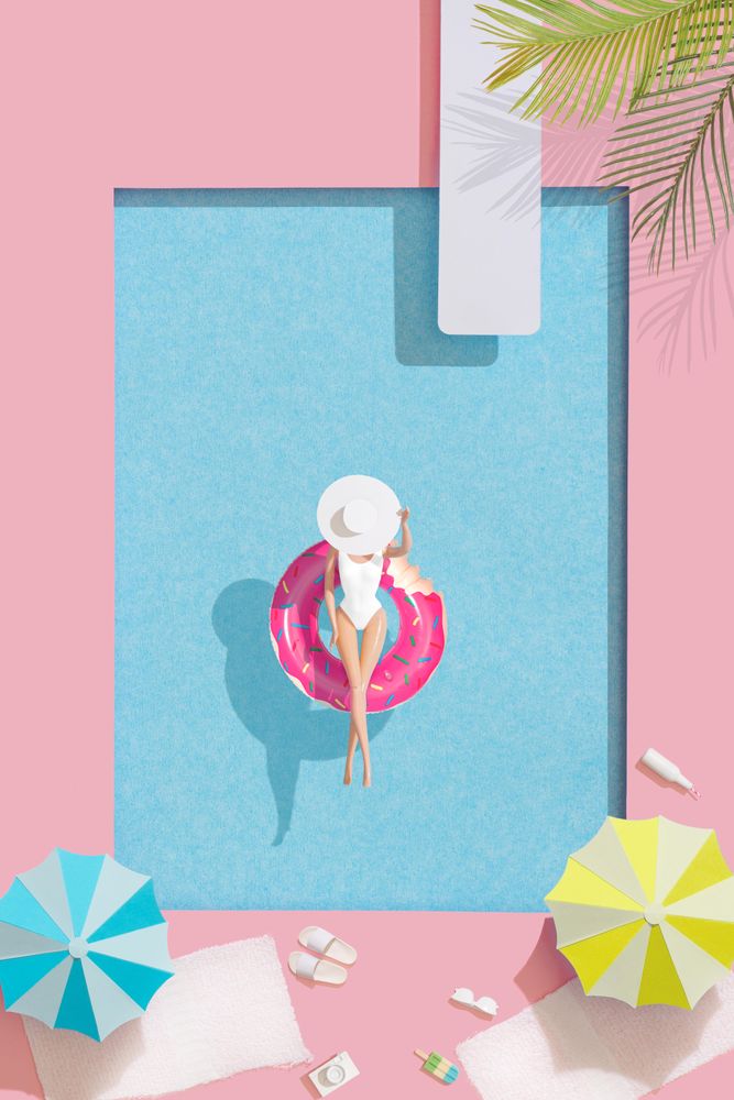 Barbie-like doll in donut ring in pool, with poolside parasols and towels