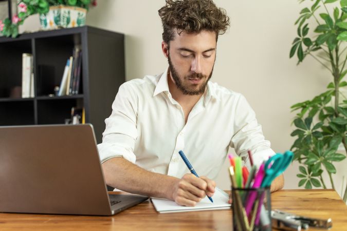 Focused young man writing in a notebook while studying at his desk.