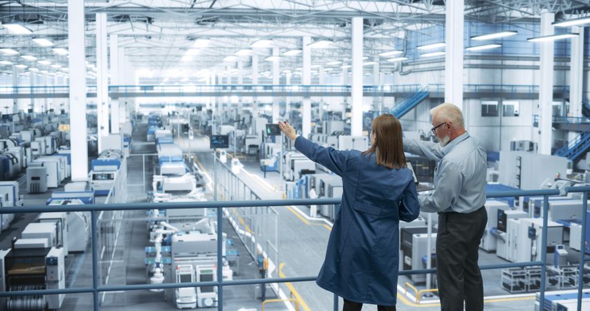 Male Engineer and Female Project Manager Standing on a Platform, Talking, Using Laptop Computer and Monitoring Production at a Modern Automated Electronics Manufacture with AI Robotic Arms