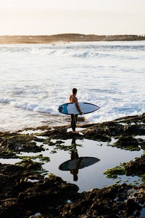 Male holding surfboard with reflection in quiet water next to ocean