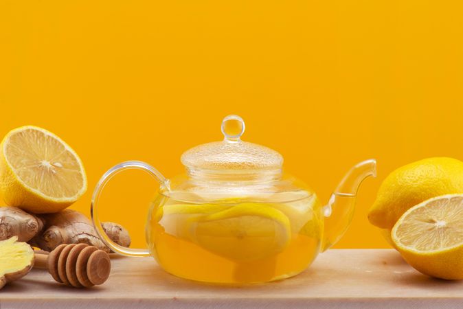 Clear pot of tea with lemon slices, surrounded by lemon halves and ginger