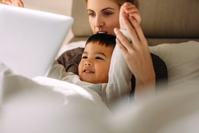 Little boy with mother enjoying watching cartoon movie on laptop while lying in bed