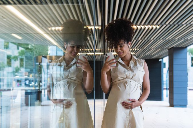 Smiling Black pregnant woman using smartphone and wireless earphones