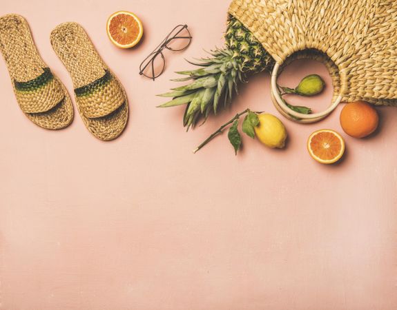 Sandals, glasses, pineapple and fruits in wicker bag, on pink background, with copy space