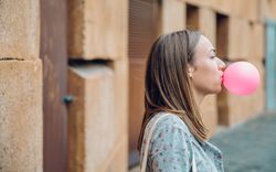 Side view of female standing outside in front of stone wall blowing bubble with pink gum with copy space