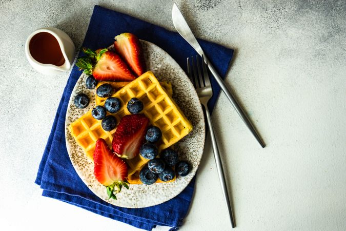 Top view of waffle breakfast with blueberries & strawberries served on grey counter with blue napkin