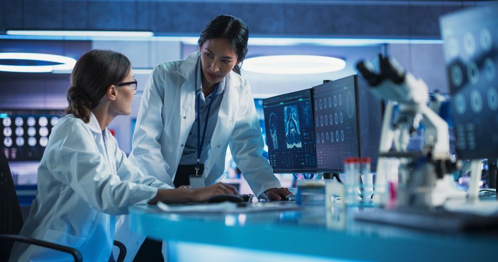 Medical Hospital Research Laboratory: White And Asian Female Scientists Talking And Using Desktop Computer With MRI Scans Of Human Brain.