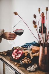Rustic setting of women holding wine, with cheese, long candles, pomegranate, and dried poppies