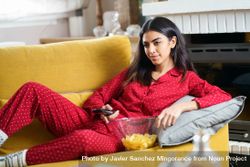 Female in red pajamas relaxing on sofa with bowl of chips and remote control 0vZRR0