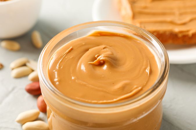 Glass jar with peanut butter, peanut, cup of coffee and peanut butter sandwich on grey background, closeup and space for text